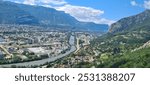 A breathtaking view of Grenoble from a high vantage point in the mountains, capturing a river winding through the city, with charming houses nestled between the flowing river and the mountains