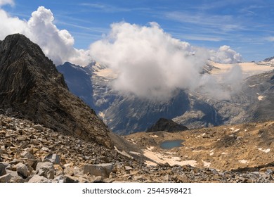 A breathtaking view of a high-altitude mountain landscape featuring a small glacial lake, rugged terrain, and clouds rolling over the peaks, set against a vibrant blue sky - Powered by Shutterstock