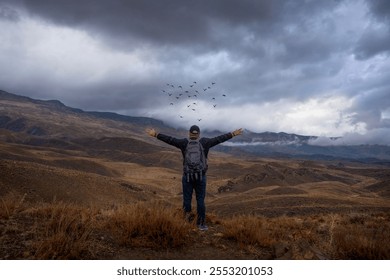 A breathtaking view of a hiker standing on a hilltop with arms outstretched, embracing the beauty of a dramatic mountainous landscape under a cloudy sky. Birds are seen flying in the distance. - Powered by Shutterstock