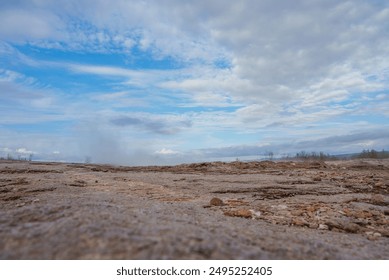 A breathtaking view of Iceland's geothermal region featuring a rocky terrain, a plume of steam from a geyser or hot spring, and distant mountains under a partly cloudy sky. - Powered by Shutterstock