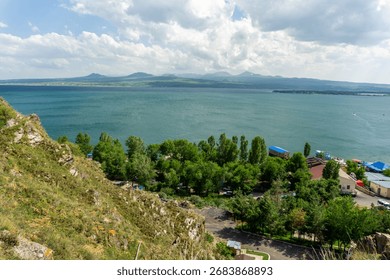 Breathtaking view of Lake Sevan in Armenia, seen from a rocky hillside with lush green trees, mountains in the distance, and vibrant lakeside buildings under a partly cloudy sky