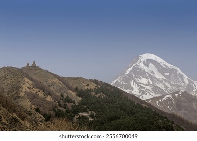 A breathtaking view of Mount Kazbek in Georgia, with its snow-capped peak rising majestically under a clear blue sky. A historic church sits atop a green ridge, surrounded by forests - Powered by Shutterstock