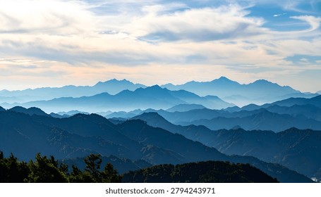A breathtaking view of multiple mountain ranges fading into the distance, shrouded in a soft blue haze under a dynamic sky with scattered clouds.