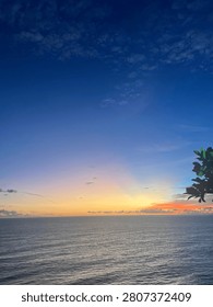 A breathtaking view of the open ocean under a expansive sky at dusk, with soft clouds catching the golden and orange glow of the sun sinking below the horizon line