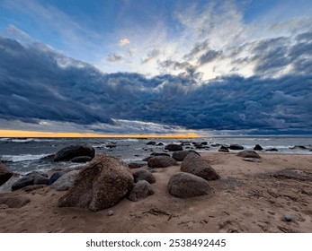 A breathtaking view of a rocky beach at sunset, with waves crashing against the stones under a dramatic sky. Dark clouds stretch across the horizon, while the setting sun casts a warm golden glow - Powered by Shutterstock