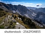 A breathtaking view of rugged mountains with a clear blue sky. The landscape features rocky terrain, lush greenery, and a distant view of a quarry or mining area