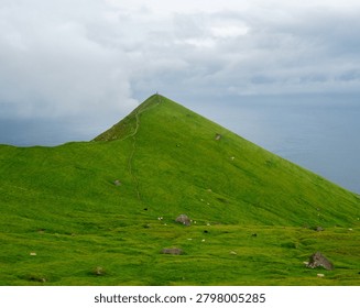 A breathtaking view of a sharp, cone-shaped green mountain peak under a dramatic cloudy sky in the Faroe Islands. A narrow dirt path winds up the steep, lush ridge toward the summit, while small sheep graze on the vast grassy slopes overlooking the calm misty ocean.