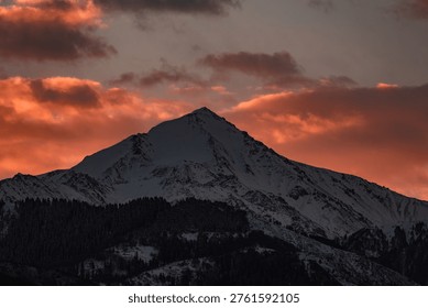 A breathtaking view of a sharp, snow-covered mountain peak silhouetted against a dramatic sunset sky. Glowing orange and pink clouds create a fiery backdrop for this serene winter alpine landscape, Almaty, Kazakhstan, Central Asia, Satpayev peak, Soviet Peak