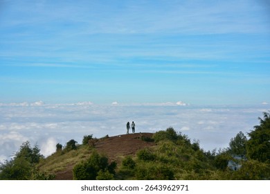 A breathtaking view from the top of a mountain as two hikers stand hand in hand above the clouds, surrounded by untouched nature and a brilliant blue sky.