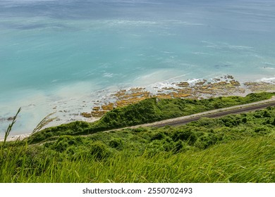 A breathtaking view of turquoise coastal waters, rocky shoreline, and railway tracks winding through lush green cliffs under a sunny sky - Powered by Shutterstock