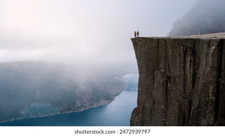 A breathtaking view of two figures standing on the edge of a towering cliff in Norway, overlooking a misty valley and a deep blue fjord. a couple of men and women hiking the Preikestolen, Norway - Powered by Shutterstock