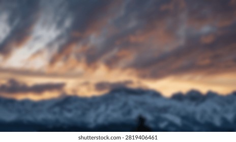 A breathtaking view of a vibrant sunset casting warm hues across a cloudy sky above majestic snow-covered mountains. A solitary person stands in silhouette, observing the scene.