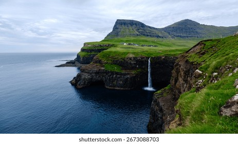 A breathtaking waterfall flows down rugged cliffs into the serene waters of the Atlantic Ocean, surrounded by vibrant green hills in the Faroe Islands, Mulafossur waterfall near Gasadalur village