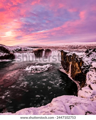Breathtaking winter landscape with a majestic waterfall amidst snow-covered rocks under a vibrant sunset sky. A serene, tranquil scene capturing nature’s grandeur in an icy wilderness.”
