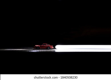 Brecht, Belgium - Februari 21 2020: A Red Toy Car Driving Over A Wooden Table In The Darkness With A White Light Painted Light Trail Behind It, The Head Lights Of The Car Are Also Light Painted.