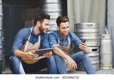 Brewery, Tech, Online Ordering And Sales Of Products, Successful Family Business. Rest And Break. Happy Attractive Young Men In Aprons Sit In Warehouse With Metal Kegs And Look At Phone And Tablet