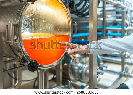 Brewery worker checking the vibrant orange liquid inside a stainless steel fermentation tank with a clear viewing window, observing the brewing process in a modern industrial beer factory