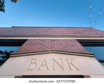 Brick Building Facade with the Word "BANK" Engraved in Stone. Modern brick building with a bank sign on the front. View from below. Clear blue sky above with string lights hanging from the building. - Powered by Shutterstock