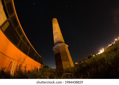 Brick Chimney Of Old Abandoned Factory Next To Abandoned Orange Bus, Night Photo With Starry Sky