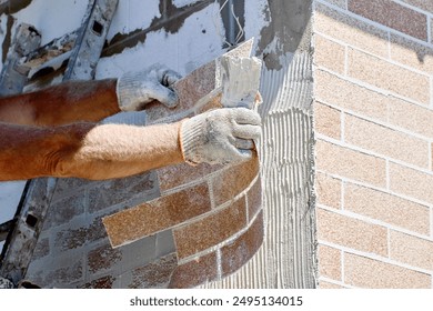 Brick cladding, also called brick veneer is attached to exterior of house made of aerated concrete blocks, construction worker uses tile adhesive.