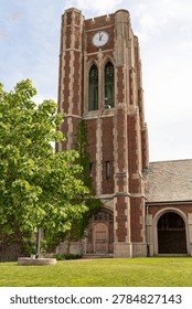 A brick clock tower with a large circular clock face at the top. The structure features tall arched windows and ornate stone detailing. Ivy climbs partially up the tower's facade