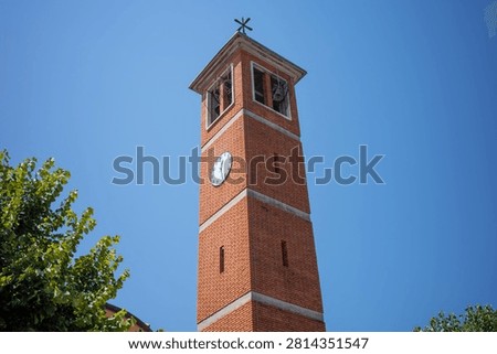 A brick clock tower is located in the town square under a clear blue sky. The tower has a clock on its front and greenery around its base.