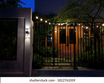 Brick Color House Has A Small Garden In Front Of The House Before Reaching The Metal Door, Leaving A Gap Inside And Hanging A Light Bulb At The Top Of The Door