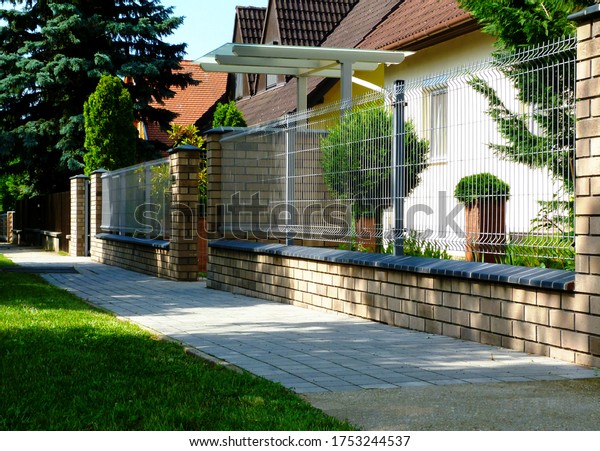 Brick fence post pier with ceramic cap stone.\
detail with painted wire mesh grille fence panels. house in the\
background with beautiful front yard. shrubs and plants. home\
ownership & security\
concept