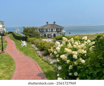 A brick pathway lined with blooming hydrangeas leads to a charming house overlooking a peaceful waterfront with boats and a serene sky in the background. Perfect summer coastal setting.