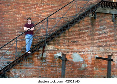 Brick wall and fire escape serves as a resting place for young teen.  He is wearing jeans with hole at knee and a torn burgundy tee shirt.