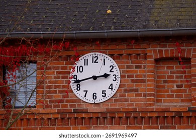 A brick wall with a round clock mounted on it, part of a window and branches with red leaves are also visible.