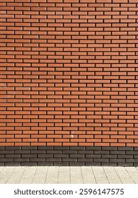 A brick wall with a uniform pattern of reddish-brown bricks. The bricks are neatly aligned in horizontal rows. At the base of the wall, there's a section of a pavement or sidewalk made of square tiles