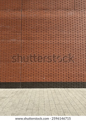 A brick wall with a uniform pattern of reddish-brown bricks. The bricks are neatly aligned in horizontal rows. At the base of the wall, there's a section of a pavement or sidewalk made of square tiles