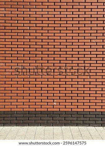 A brick wall with a uniform pattern of reddish-brown bricks. The bricks are neatly aligned in horizontal rows. At the base of the wall, there's a section of a pavement or sidewalk made of square tiles