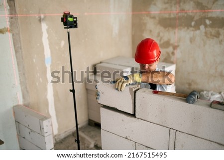 Bricklayer or mason lays bricks to construct wall of autoclaved aerated concrete blocks. Brickwork worker contractor doing precise masonry of foam concrete with spirit and laser levels.