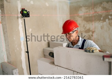 Bricklayer using rubber mallet hammer to tap and level concrete blocks wall. Contractor uses precise spirit bubble and laser level for brickwork. Worker constructs a wall in new apartment real estate.