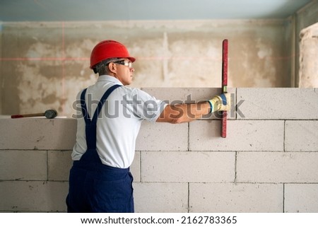 Bricklayer using spirit bubble and laser level to precise check concrete blocks on wall. Contractor uses tools for brickwork. Worker constructs a wall in new apartment real estate.
