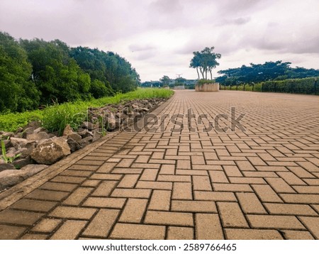 Brick-Paved Path with Lush Green Trees and Overcast Sky. A peaceful open path featuring brick paving, greenery, rocks, and a cloudy atmosphere.