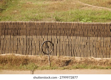 Bricks neatly stacked in rows form a long, sturdy wall. Single tire rests on a pole beside the wall. Sunlight bathes the earthy tones of the bricks. Grass grows wild around the base