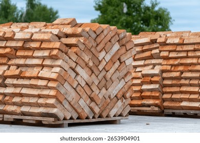 Bricks are neatly stacked in a zigzag pattern at a construction site surrounded by greenery, indicating preparation for building.