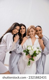 bridal party, four women, joyful bride holding bouquet with white flowers near bridesmaids in silk robes, cultural diversity, togetherness, friendship goals, brunette and blonde women