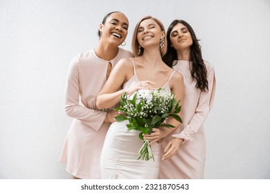 bridal party, special occasion, brunette and interracial bridesmaids hugging blonde bride, friendship goals, grey background, happy multicultural girlfriends, cultural diversity, bridal bouquet - Powered by Shutterstock