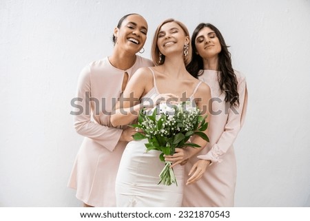 bridal party, special occasion, brunette and interracial bridesmaids hugging blonde bride, friendship goals, grey background, happy multicultural girlfriends, cultural diversity, bridal bouquet