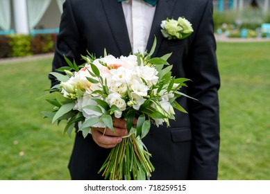 Bridal wedding bouquet of white flowers and greenery with ribbons in groom hand. Groom waiting for bride outdoor. Young handsome groom in black suit standing and holding wedding bouquet