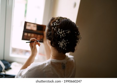 Bride applies makeup while standing by the window in a hotel room before the wedding ceremony, back view 