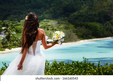 Bride, Beautiful Young Girl With Dark Hair In White Wedding Dress With Bouquet On Background Of Beach With Blue Water. Seychelles