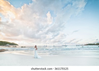 Bride, Beautiful Young Girl With Dark Hair In White Wedding Dress With Bouquet On Background Of Beach With Blue Water. Seychelles