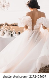 A Bride With Black Hair In A White Airy Dress With An Open Back And A Flowing Train. Standing With His Back Against The Background Of A Banquet Table With A Wedding Decor.