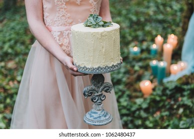 Bride In Blush Wedding Dress Holding An Unusual Wedding Cake Decorated With Succulent Plant With Forest Floor In The Background