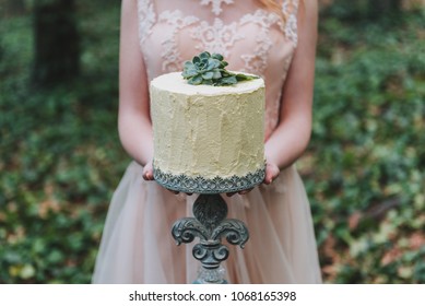 Bride In Blush Wedding Dress Holding An Unusual Wedding Cake Decorated With Succulent Plant With Forest Floor In The Background